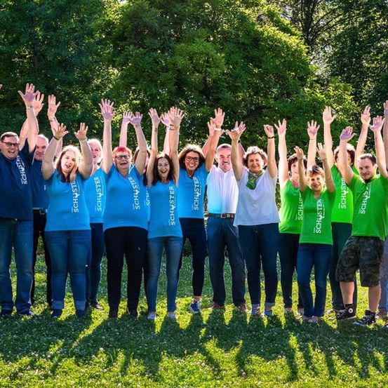 Eine Gruppe von Menschen in blauen und grünen Shirts, die in einem Park stehen und für ein Foto mit erhobenen Händen posieren.
