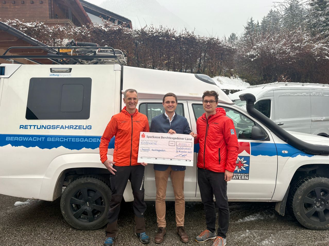Drei Männer stehen vor einem weiß-blauen Fahrzeug, einer hält einen großen Scheck. Sie tragen alle Jacken und lächeln. Dahinter befindet sich ein verschneiter Bereich mit einem Gebäude.