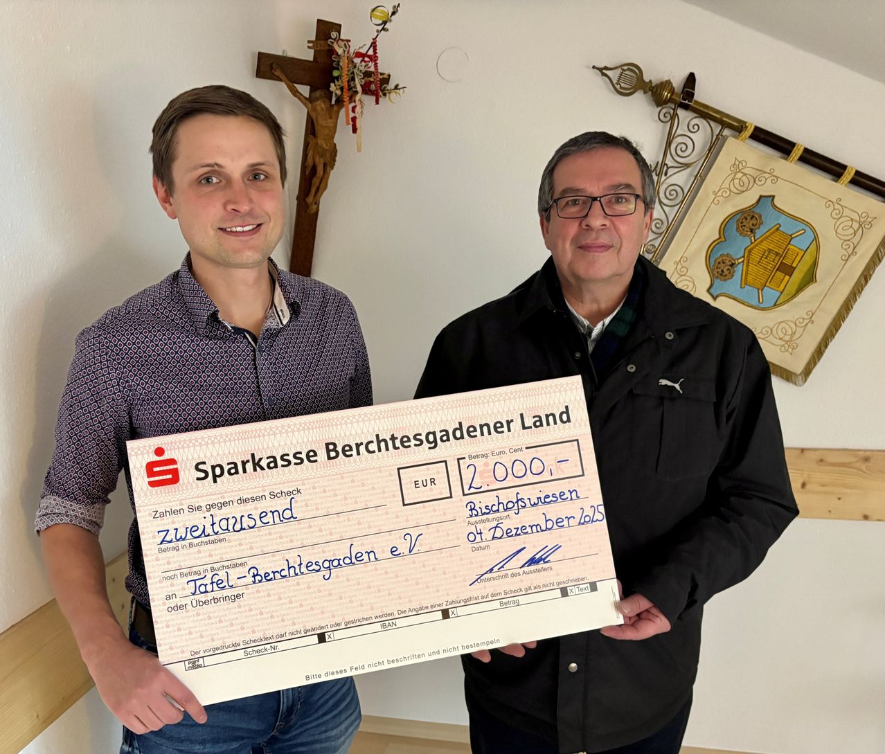 Zwei Männer stehen und halten einen Scheck in der Hand, einer der Männer lächelt. Der Scheck hat einen Nennwert von 2.000 Euro. Der Mann auf der rechten Seite trägt eine Brille und eine Jacke. Hinter ihnen befindet sich eine weiße Wand mit einem Kruzifix und einer Flagge.