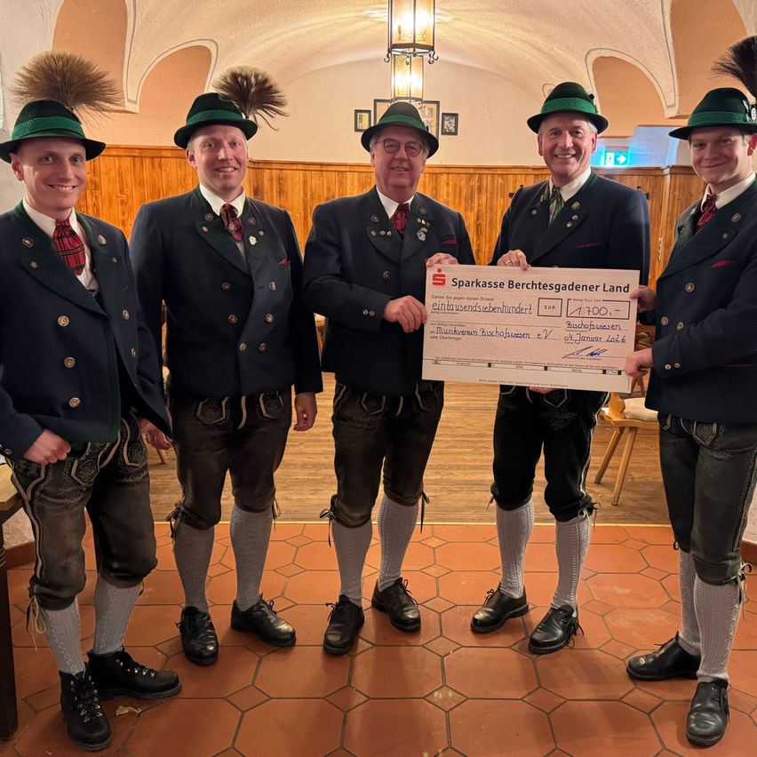 Fünf Männer in traditioneller deutscher Kleidung stehen zusammen und halten einen großen Scheck. Der Mann in der Mitte lächelt und hält den Scheck hoch. Die anderen Männer schauen interessiert zu.