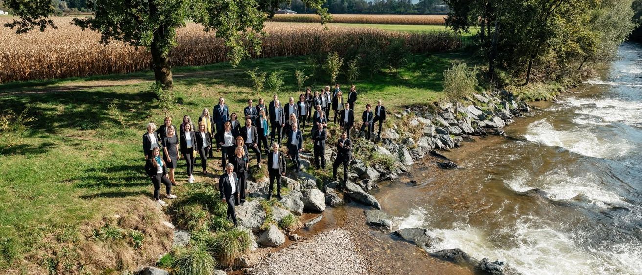 Eine Gruppe formell gekleideter Personen, die am Ufer eines Flusses steht, mit einem Maisfeld und Bäumen im Hintergrund.