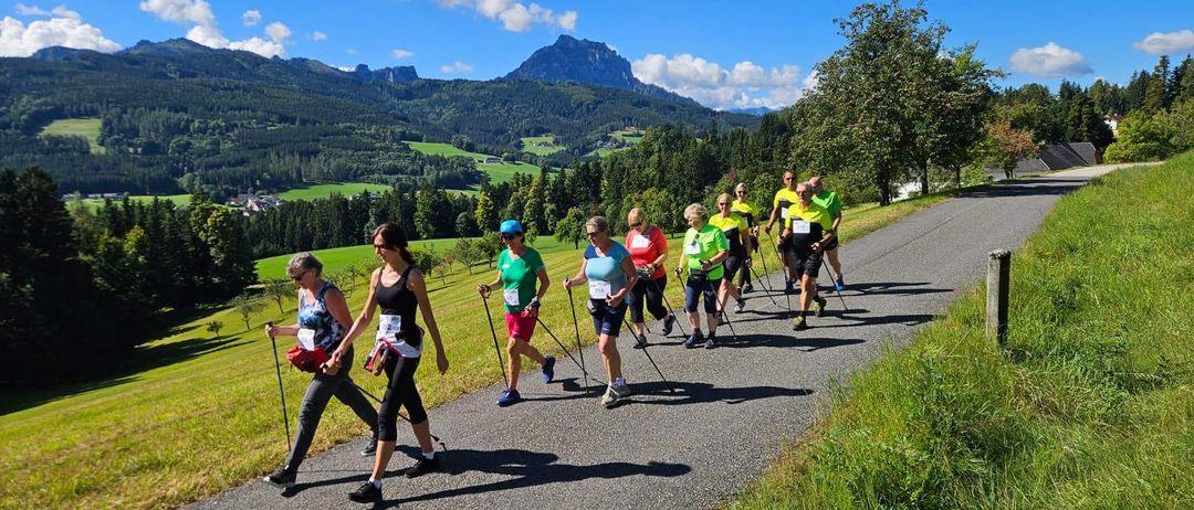 Eine Gruppe von Menschen läuft mit Trekkingstöcken auf einem Schotterweg mit grasbewachsener Seite. Sie tragen Turnschuhe und Rucksäcke. Hinter ihnen stehen Bäume, Gras und Berge.