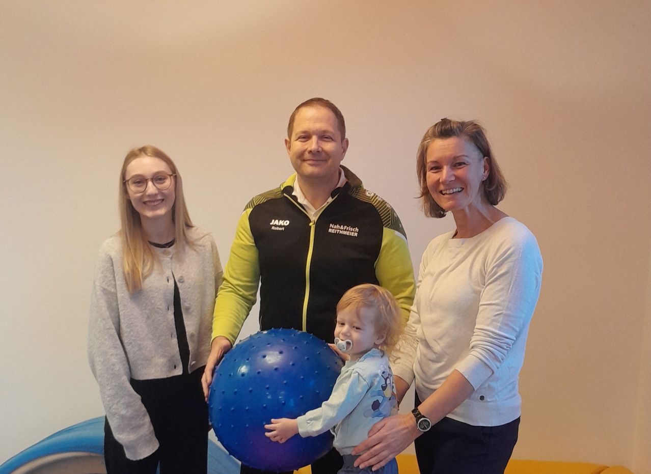Drei Personen, darunter ein Mann und zwei Frauen, stehen mit einem Kleinkind vor einer weißen Wand. Der Mann hält einen blauen Ball und lächelt. Das Kleinkind trägt ein weißes Shirt mit einem Aufdruck.