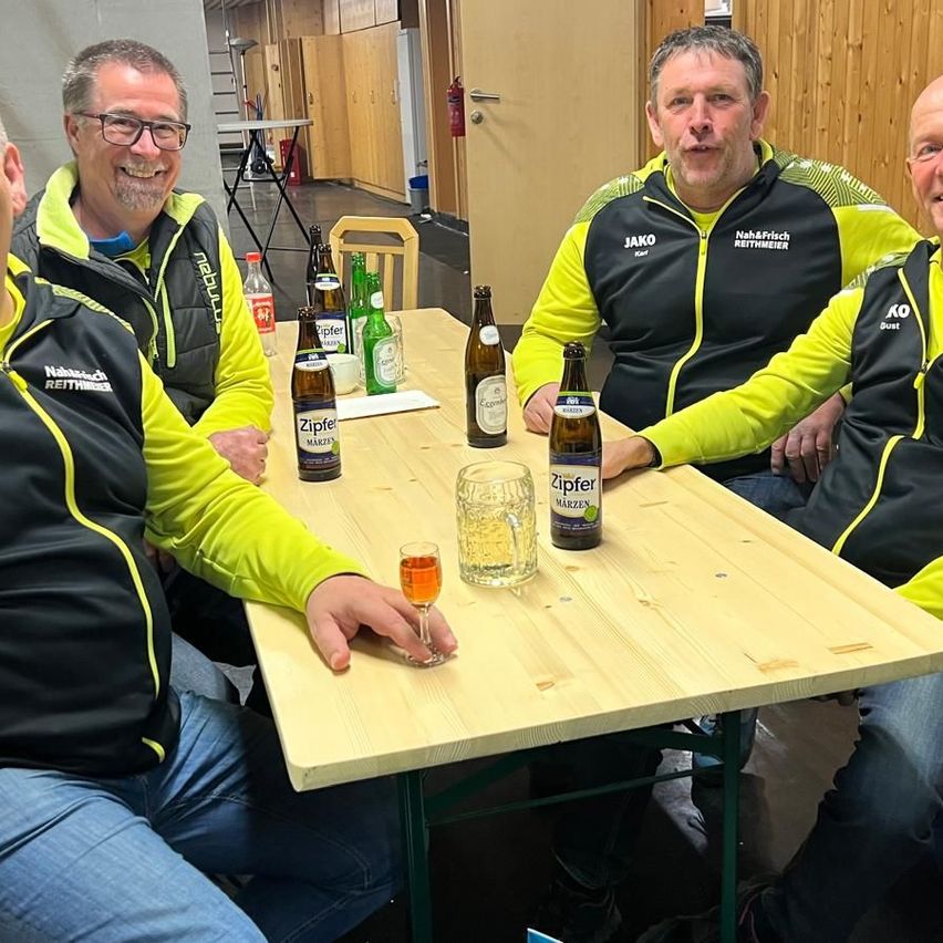 Four men in matching yellow and black jackets sit around a table with bottles and a glass of beer. One man holds a glass.