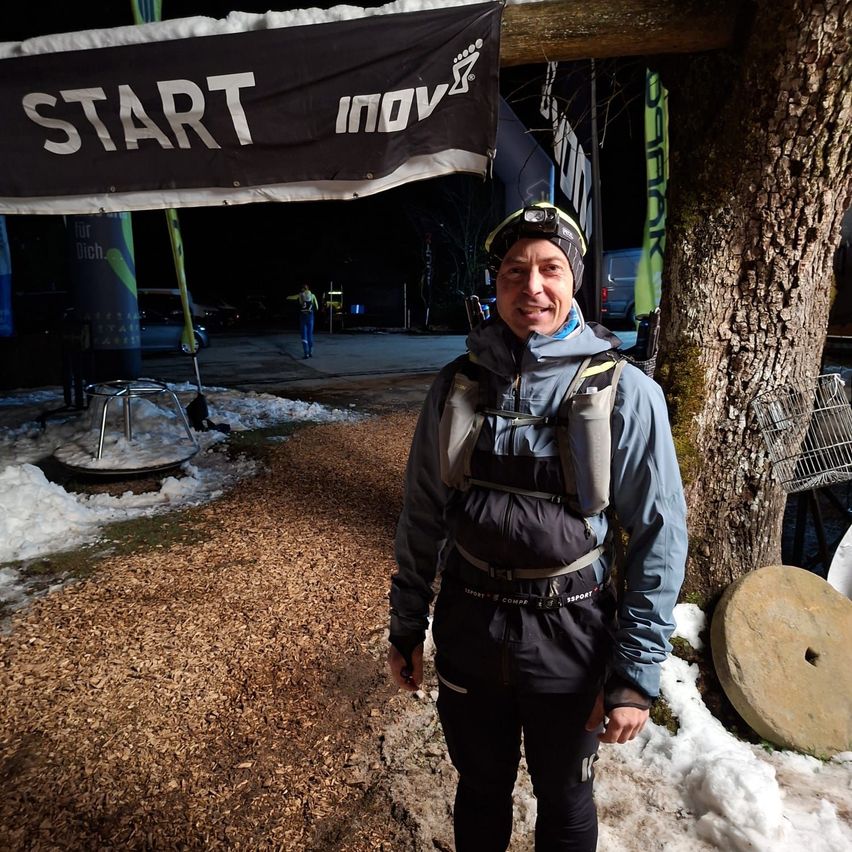 A man stands under a black banner with the word 'Start' on a snowy path. He is smiling and wearing a winter jacket. Behind him, a tree trunk and a metal wheel are visible.