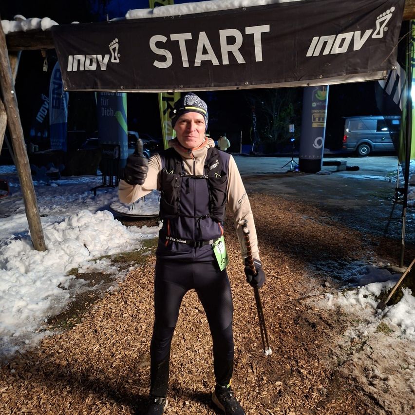 A man dressed in winter gear stands under a sign that says Start, holding a ski pole and giving a thumbs-up gesture.