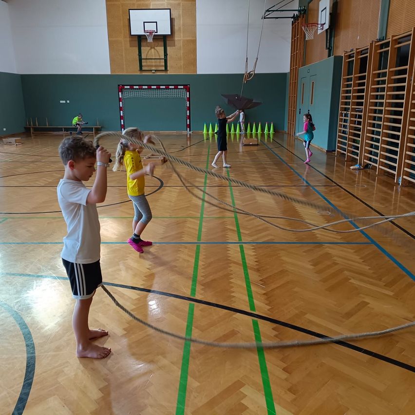 Kinder spielen mit Seilen in einer Sporthalle. Ein Mädchen hält ein Seil, während ein anderes läuft. Ein Mädchen in Blau geht in der Nähe.