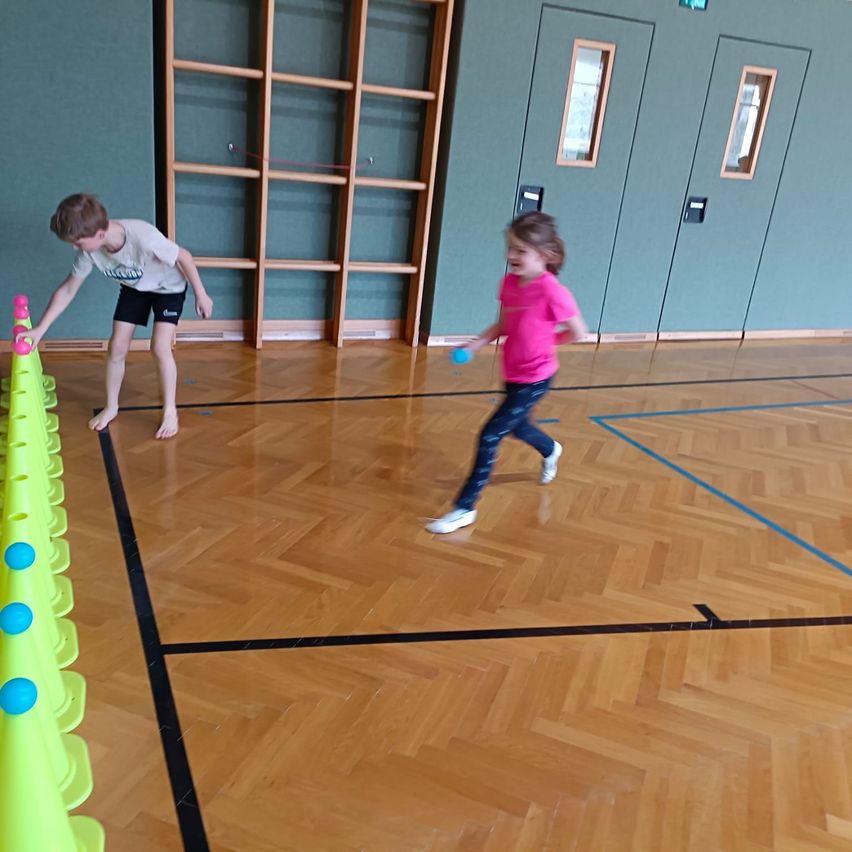 Zwei Kinder spielen in einer Turnhalle, rennen um Kegel und halten Bälle. Der Boden ist hölzern mit blauen Linien.
