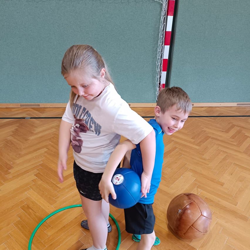 Ein junges Mädchen und ein Junge spielen in einer Turnhalle mit einem blauen Ball und einem braunen Fußball. Das Mädchen hält die Schulter des Jungen, während sie sich gegenseitig ansehen. Ein grüner Hula-Hoop liegt in der Nähe.