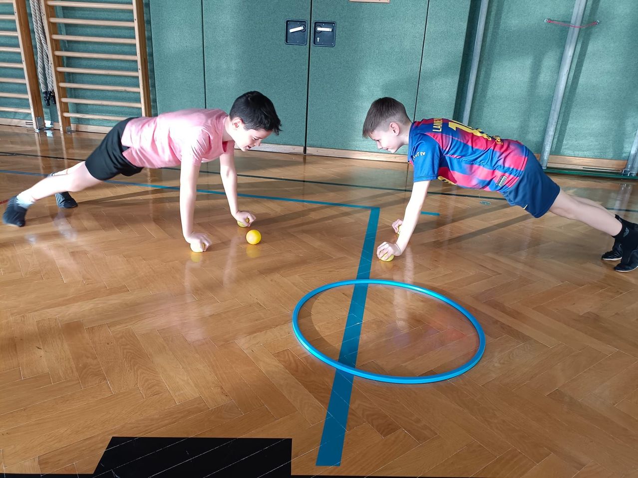 Zwei Jungen machen Liegestütze auf einem Holzboden mit einem blauen Hula-Hoop in der Nähe. Beide tragen Sporttrikots und halten gelbe Bälle.