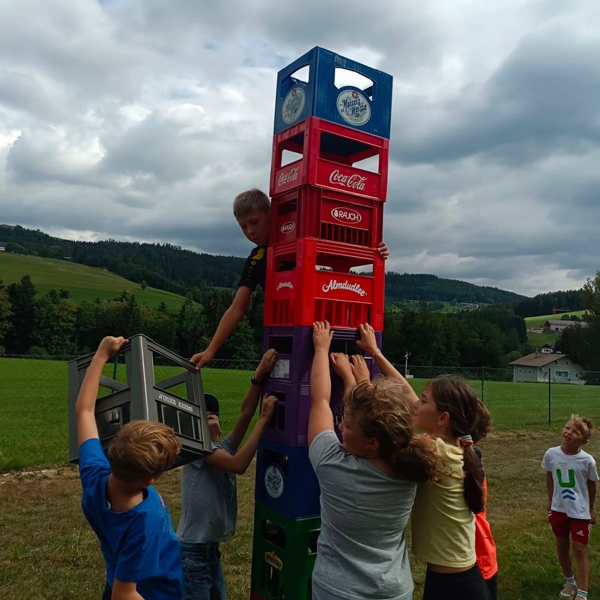 Eine Gruppe von Kindern klettert auf einen hohen Stapel bunter Kisten im Freien. Die Kisten sind in verschiedenen Farben gestapelt, darunter rot, blau und grün. Die Kinder halten sich an den Kisten fest, und ein Kind ist oben. Im Hintergrund befindet sich ein Grasfeld mit Bäumen und Bergen.