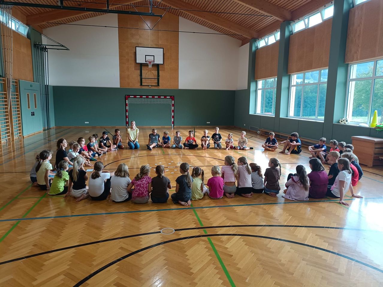 Eine Gruppe von Kindern und einer Frau sitzen im Kreis auf einem Basketballplatz. Die Frau steht in der Mitte. Der Platz hat einen Basketballkorb und ein Tor. Die Wände sind grün, und es gibt Fenster mit Bäumen draußen.