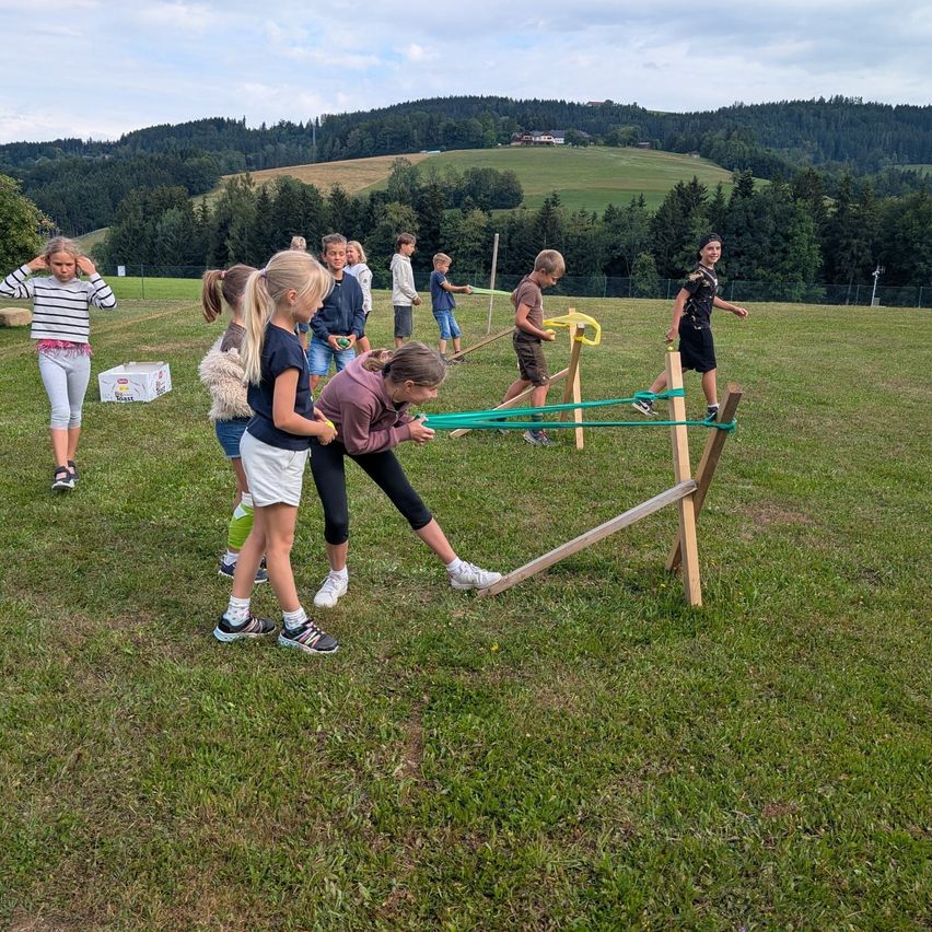 Eine Gruppe von Kindern spielt ein Spiel auf einer Wiese mit einem hölzernen Hindernisparcours und einem Berg im Hintergrund.