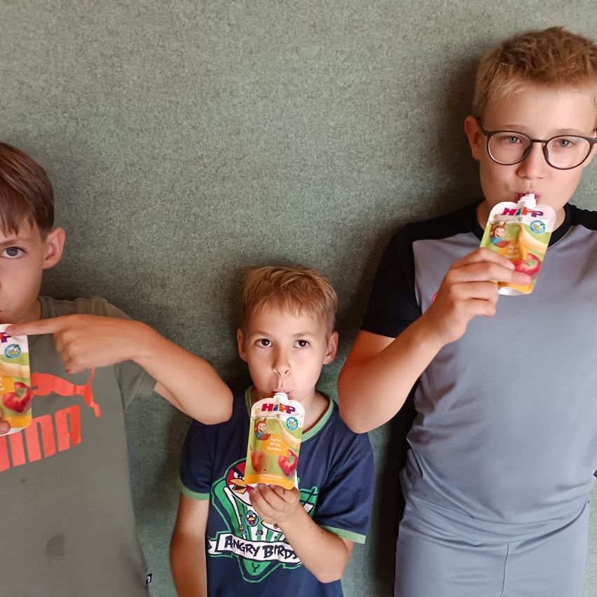 Drei Jungen stehen vor einer grünen Wand, jeder hält einen Beutel mit Fruchtsaft. Der Junge auf der linken Seite zeigt, der in der Mitte trinkt und der Junge auf der rechten Seite trinkt ebenfalls.