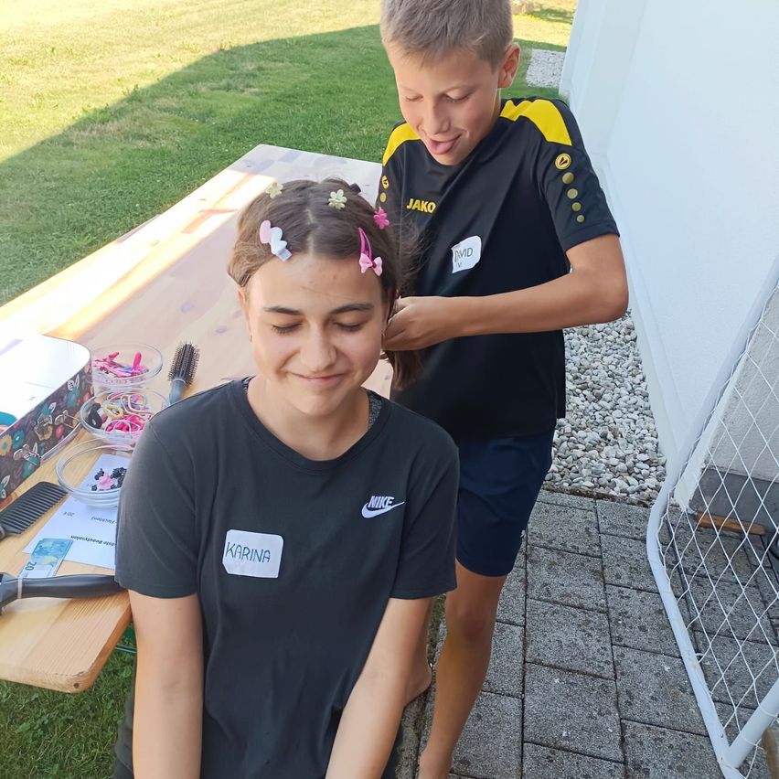 Ein junges Mädchen mit einem Pferdeschwanz sitzt auf einer Bank, während ein Junge hinter ihr steht und ihr Haar in Ordnung bringt. Sie sind draußen in der Nähe eines Tisches mit verschiedenen Haaraccessoires.