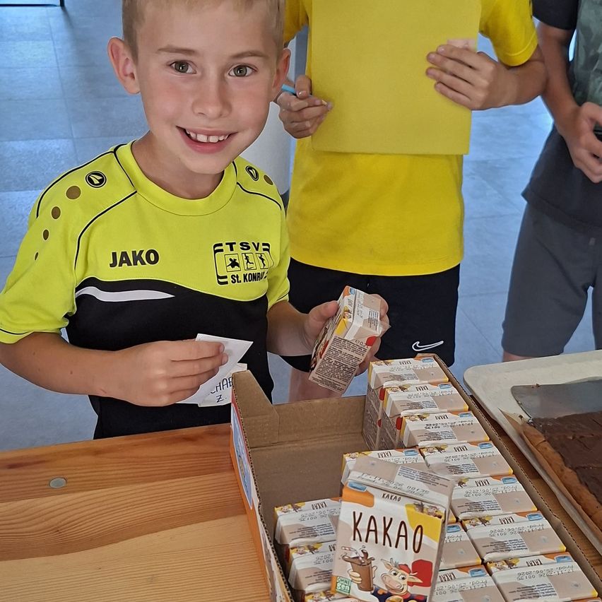 Ein junger Junge in einem gelben Sporttrikot hält eine Schachtel Kakao-Schokolade und lächelt in die Kamera. Dahinter stehen zwei Personen, eine hält ein gelbes Papier.