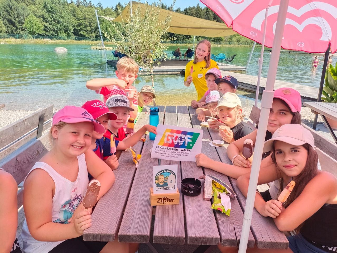 Eine Gruppe von Kindern sitzt an einem Picknicktisch am See, isst Eis und Snacks, mit einem Schild, das GWF steht, und einem Baum im Hintergrund.