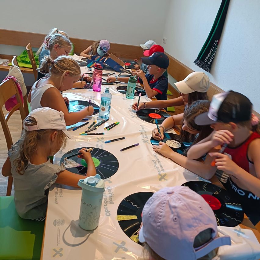 Eine Gruppe von Kindern sitzt um einen Tisch herum und malt mit Markern und Stiften auf Vinylplatten. Der Tisch ist mit einem weißen Tuch bedeckt, und jedes Kind hat eine Wasserflasche und eine Platte vor sich.