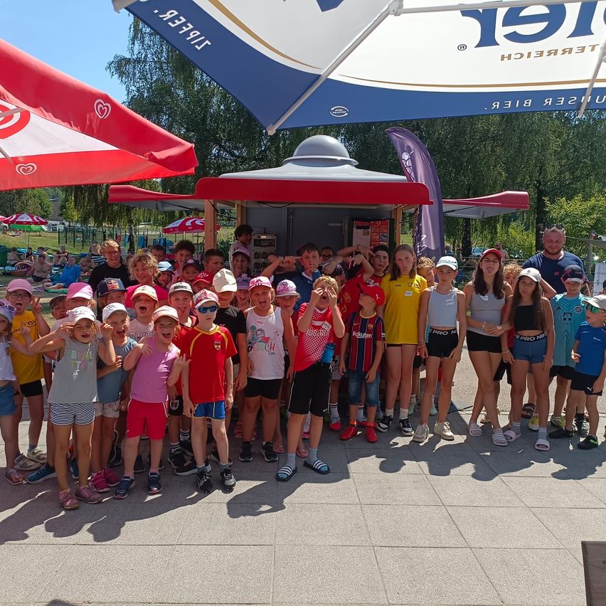 Eine Gruppe von Kindern und Erwachsenen posiert für ein Foto vor einem Eiswagen. Die Kinder tragen Mützen und Turnschuhe, und die Erwachsenen stehen hinter ihnen. Im Hintergrund befinden sich Schirme und ein Banner.