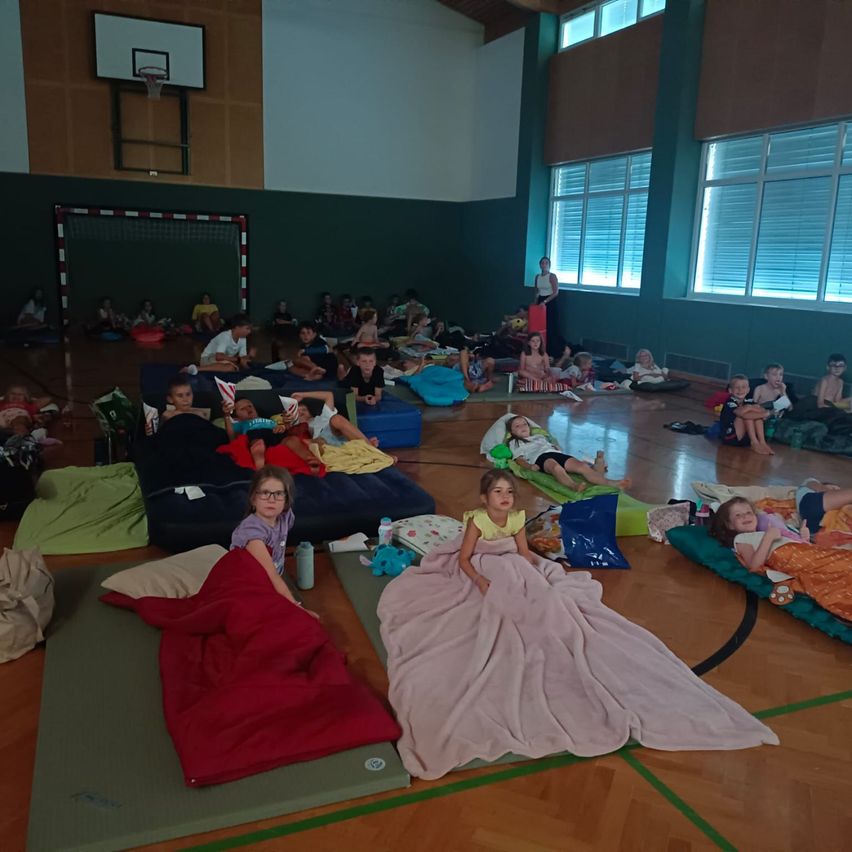 Ein Sportsaal voller Kinder, die auf Matratzen und Decken auf dem Boden schlafen, mit einem Basketballkorb im Hintergrund.
