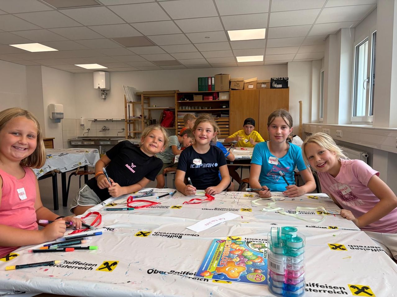 Eine Gruppe von Kindern sitzt an einem Tisch, lächelt und hält Marker in den Händen. Sie machen eine Kunstaktivität mit einem Tisch, der mit einem weißen Tuch bedeckt ist. Auf dem Tisch befinden sich verschiedene Kunstmaterialien.