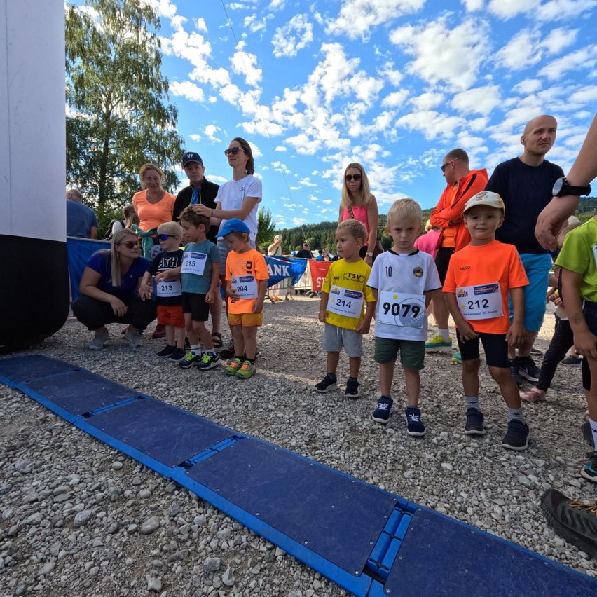 Eine Gruppe von Kindern in Sportbekleidung mit Nummern auf ihren Trikots steht in einer Reihe auf einem Schotterweg, mit Erwachsenen dahinter. Der Himmel ist blau mit verstreuten Wolken.