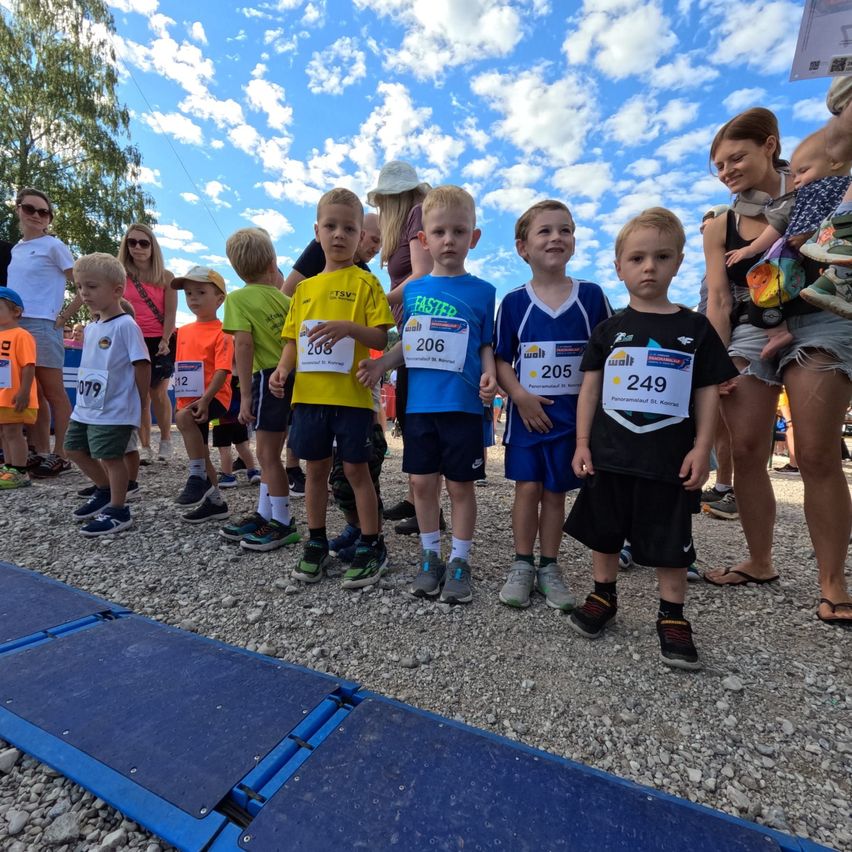 Eine Gruppe junger Jungen in nummerierten Shirts und Turnschuhen steht auf einem Kiesweg, wahrscheinlich für ein Rennen. Sie sind von anderen Kindern und Erwachsenen umgeben, von denen einige Babys tragen. Der Himmel ist blau mit einigen Wolken.