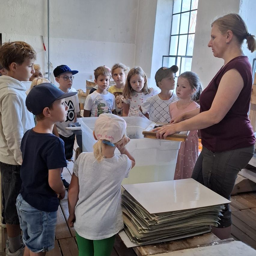 Eine Frau unterrichtet eine Gruppe von Kindern in einem Klassenzimmer und zeigt ihnen ein Holzbrett. Die Kinder stehen um einen Tisch mit einem weißen Behälter und einem Stapel weißer Papiere.
