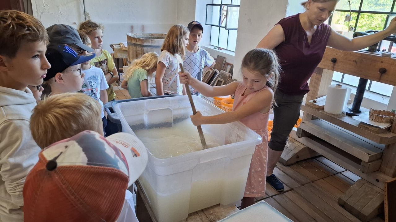 Ein kleines Mädchen rührt in einem großen Behälter mit Flüssigkeit, während eine Frau zusieht und mehrere Kinder zusehen.