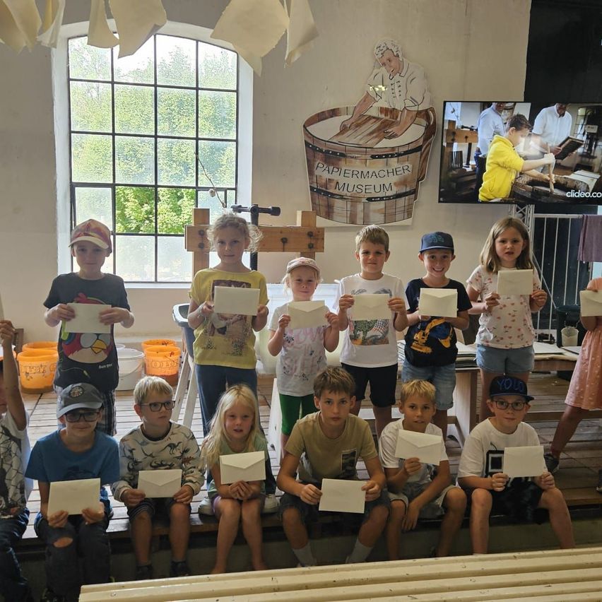 Eine Gruppe von Kindern in einem Raum, die Papiere halten, mit einem großen Bildschirm, der eine Person zeigt, die Papier herstellt.