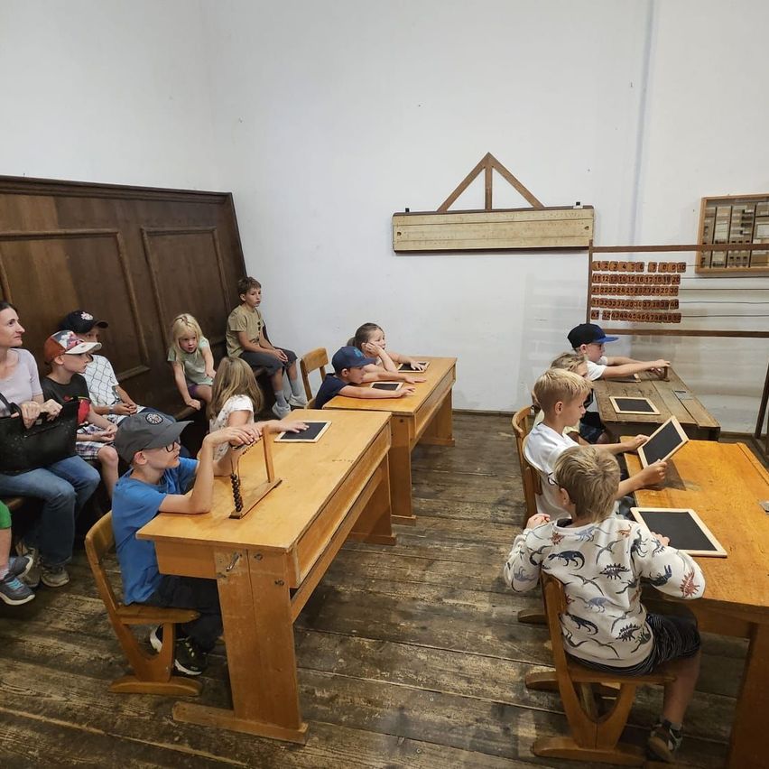 Eine Gruppe von Kindern und Erwachsenen sitzt an Holzschreibtischen in einem Klassenzimmer und benutzt Tablets. Der Raum hat Holzwände und -böden.