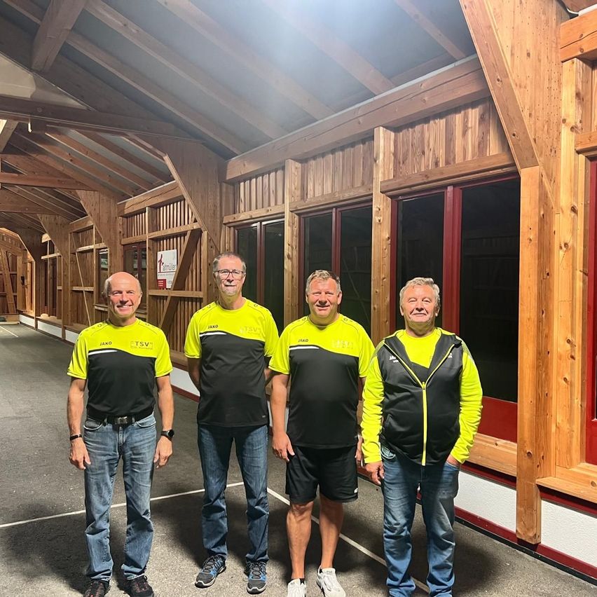 Vier Männer in passenden gelben und schwarzen Sporttrikots stehen in einem hölzernen Gebäude mit roten Fenstern. Sie lächeln und scheinen für ein Foto zu posieren.