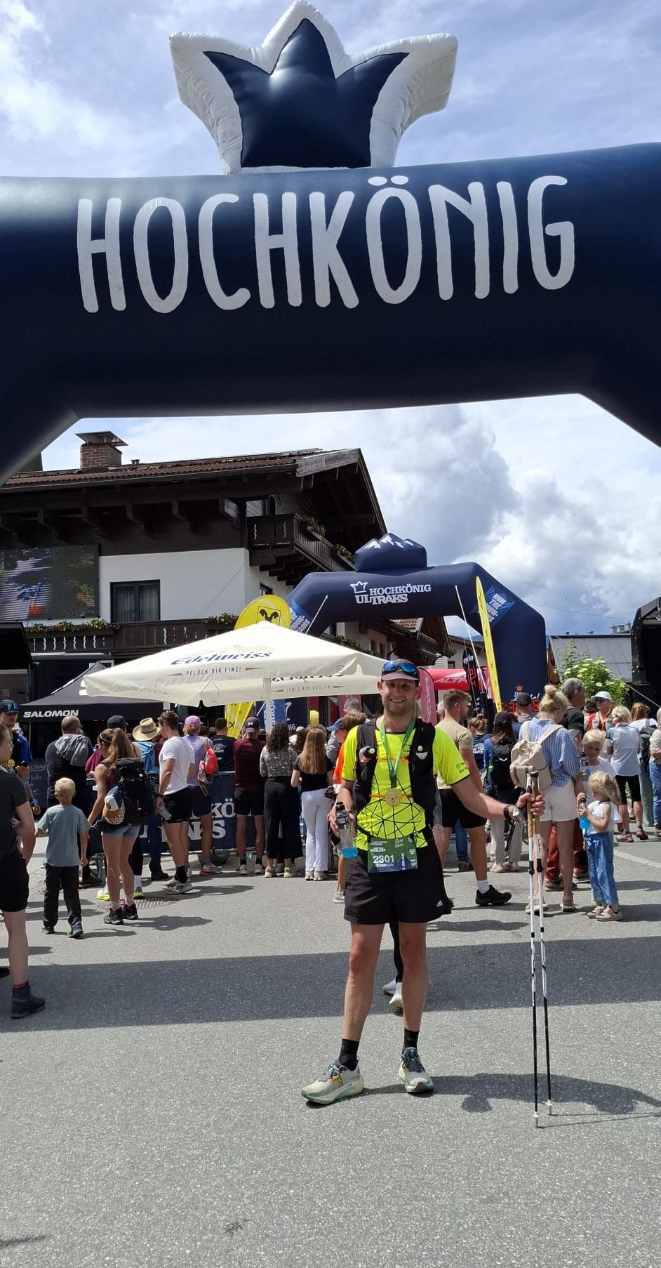 Ein Mann steht vor einer Menschenmenge unter einem Bogen, trägt eine Medaille, hält eine Wasserflasche und Trekkingstöcke. Im Hintergrund hat ein Gebäude Pflanzen auf dem Balkon.