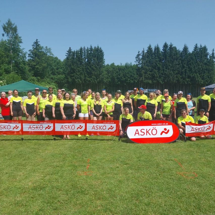 Eine Gruppe von Menschen in gelben und schwarzen Uniformen steht in einer Reihe mit einem Banner, das 'ASKO' steht. Dahinter stehen Bäume und ein grünes Zelt.