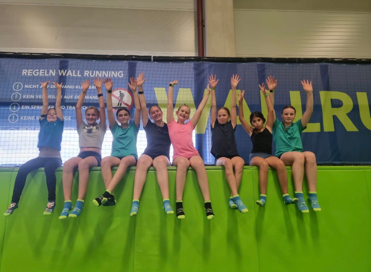 Eine Gruppe von Mädchen sitzt auf einer Wand, trägt bunte Socken und hebt ihre Hände. Sie scheinen für ein Foto zu posieren. Die Wand ist grün gestrichen. Dahinter befindet sich ein Banner mit Text.