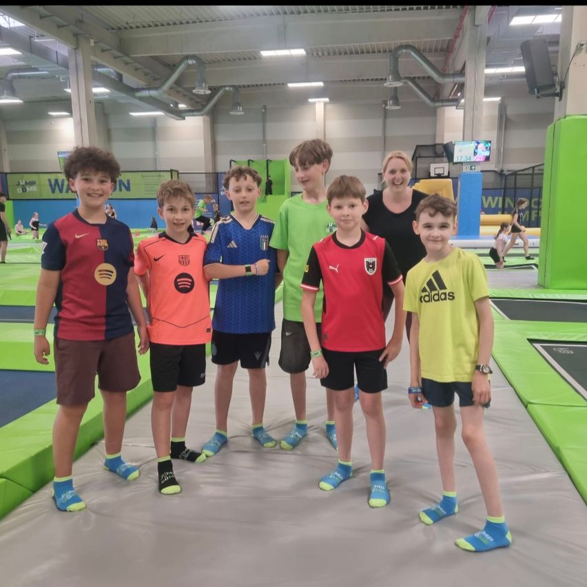 Sechs Jungen in verschiedenen Trikotfarben stehen auf einem Trampolin und lächeln für ein Foto. Eine erwachsene Frau steht hinter ihnen und lächelt ebenfalls. Der Bereich ist mit farbenfroher Ausrüstung gefüllt.