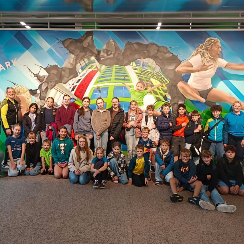 Eine Gruppe von Kindern und Erwachsenen posiert für ein Foto vor einem großen Wandbild. Das Wandbild zeigt eine springende Frau und andere Kinder in verschiedenen Posen. Die Kinder tragen Freizeitkleidung, einige mit Brille.