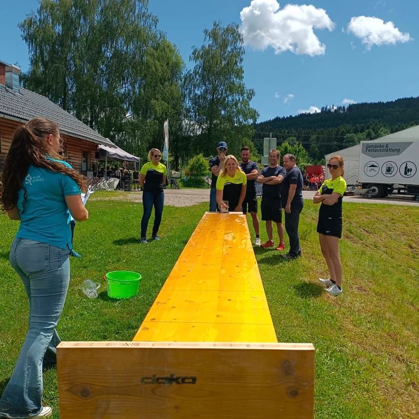 Eine Gruppe von Menschen in gleichen Uniformen steht neben einem gelben Tisch im Freien. Eine Frau steht an der Seite, während andere um den Tisch versammelt sind. Ein Gebäude, Grünpflanzen und Berge sind im Hintergrund zu sehen.