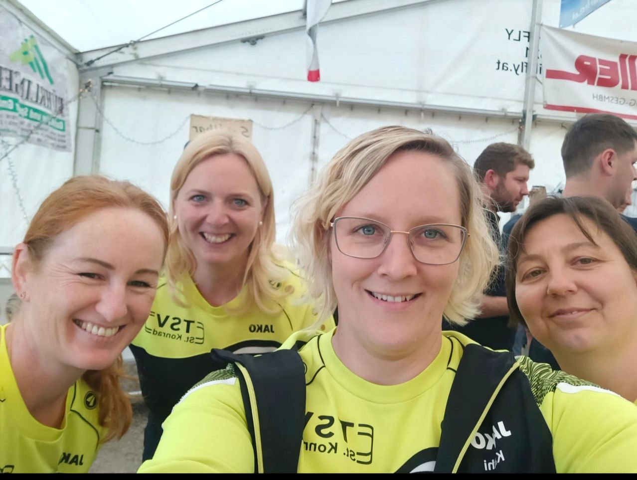 Vier Frauen in leuchtend gelben Shirts mit schwarzen und weißen Texten lächeln und posieren für ein Foto in einem Zelt.