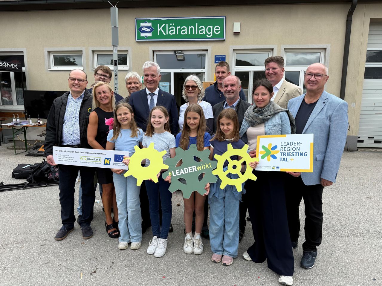 Eine Gruppe von Erwachsenen und Kindern, die Schilder halten, steht vor dem Köranlage-Gebäude. Sie scheinen alle frühlich in die Kamera zu lächeln.