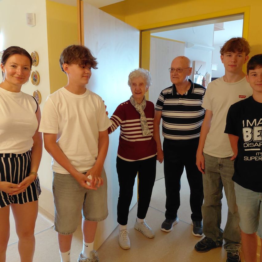 A group of people, including a young boy, an elderly woman, and a man with glasses, are standing together in a hallway. The hallway has yellow walls and a white door.