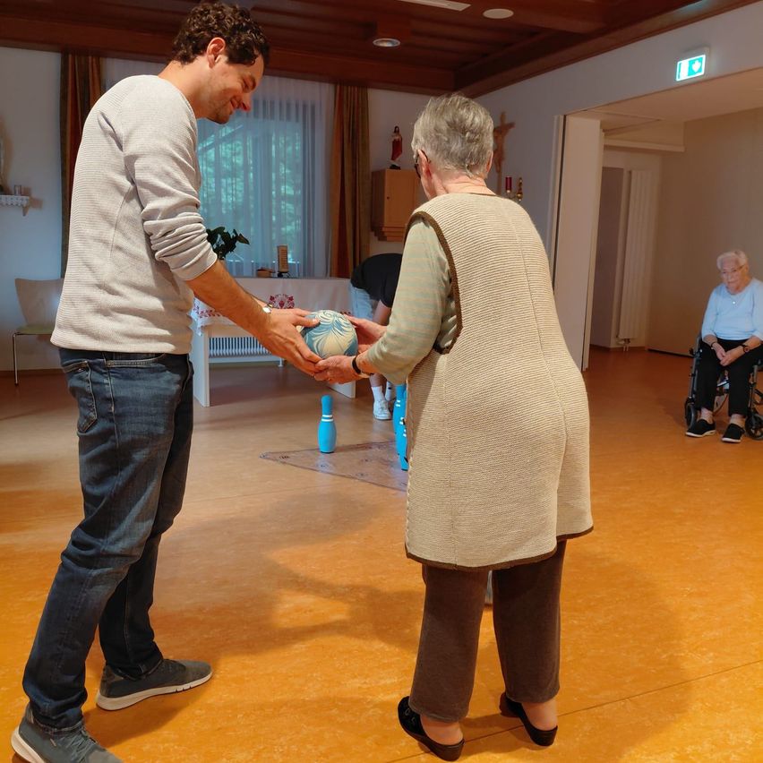 A man and an elderly woman play a game of bocce ball in a room with wooden floors and white walls.
