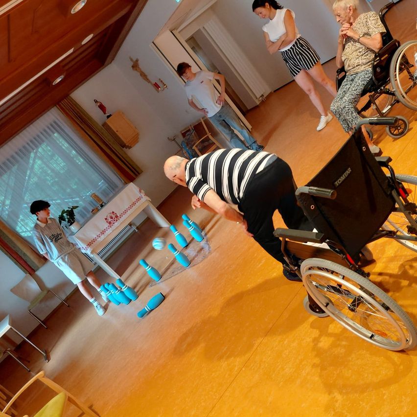 A group of people are playing a game of bowling in a room with wooden floors and white walls. An elderly man in a wheelchair is bowling, while others watch.
