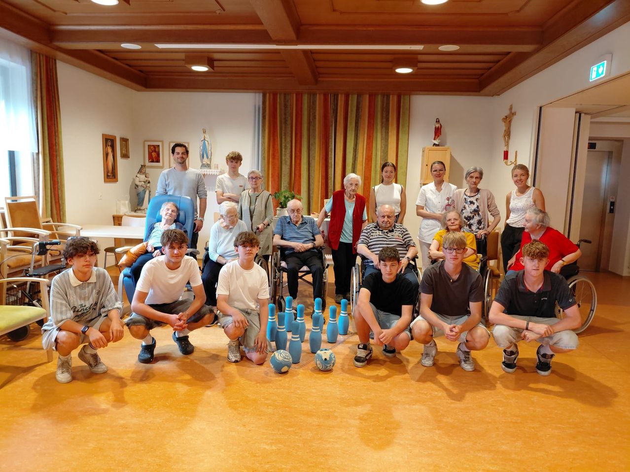 A group of people, including some in wheelchairs, are posing for a photo in a room with wooden floors and a wooden ceiling. Bowling pins and balls are on the floor.