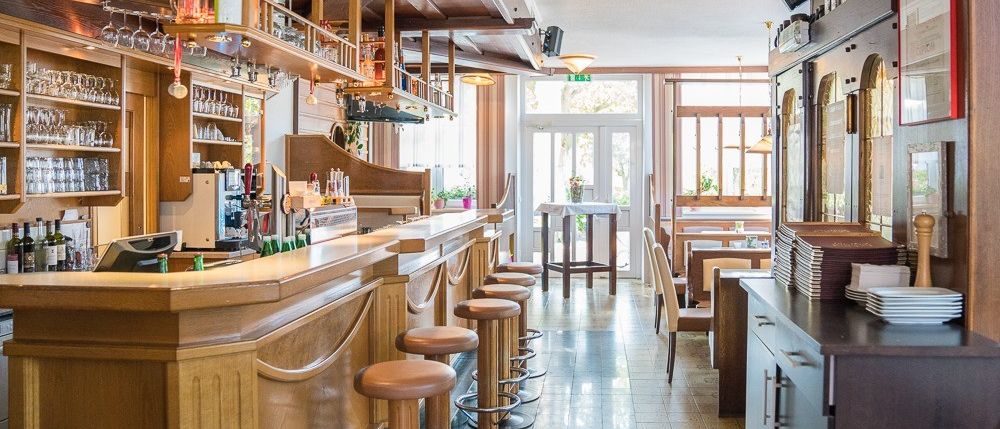 Ein Barinnenraum mit Holzregalen, Barhockern und einer Theke. Flaschen und Gläser sind auf den Regalen. Es gibt einen Tisch mit einer Vase darauf.