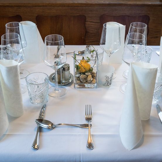 Ein Tisch mit Tischgeschirr, Gläsern, Servietten, Besteck und einer kleinen Pflanze in der Mitte. Der Tisch ist mit einer weißen Tischdecke bedeckt.