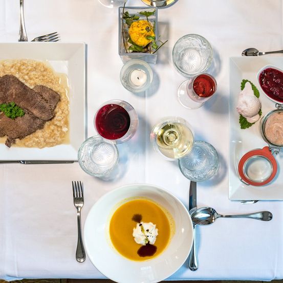 Ein weißer Tischtuch bedeckt einen Tisch mit zwei Tellern mit Essen. Der linke Teller hat ein Stück Rindfleisch und cremigen Risotto. Der rechte Teller hat eine Suppe mit einem Klecks Sahne und eine kleine Schale mit Cranberry-Sauce. Gläser und Besteck sind verstreut.
