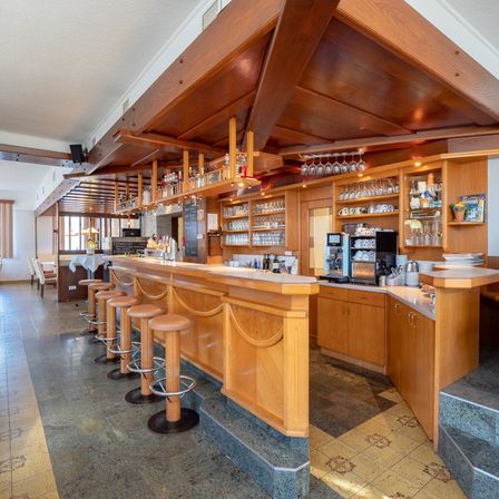 Eine Innenbar mit Holztheken, Hockern, mit Gläsern gefüllten Regalen, einer Kaffeemaschine und einer Tafel. Die Bar hat ein modernes Design mit gefliestem Boden und Decke.