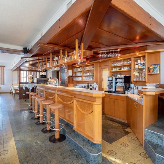 Eine Innenbar mit Holztheken, Hockern, mit Gläsern gefüllten Regalen, einer Kaffeemaschine und einer Tafel. Die Bar hat ein modernes Design mit gefliestem Boden und Decke.