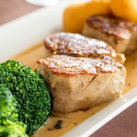 Ein Teller mit drei Stücken gebratenem Schweinekotelett in cremiger Sauce und einer Beilage aus Brokkoli-Röschen und Kartoffeln.